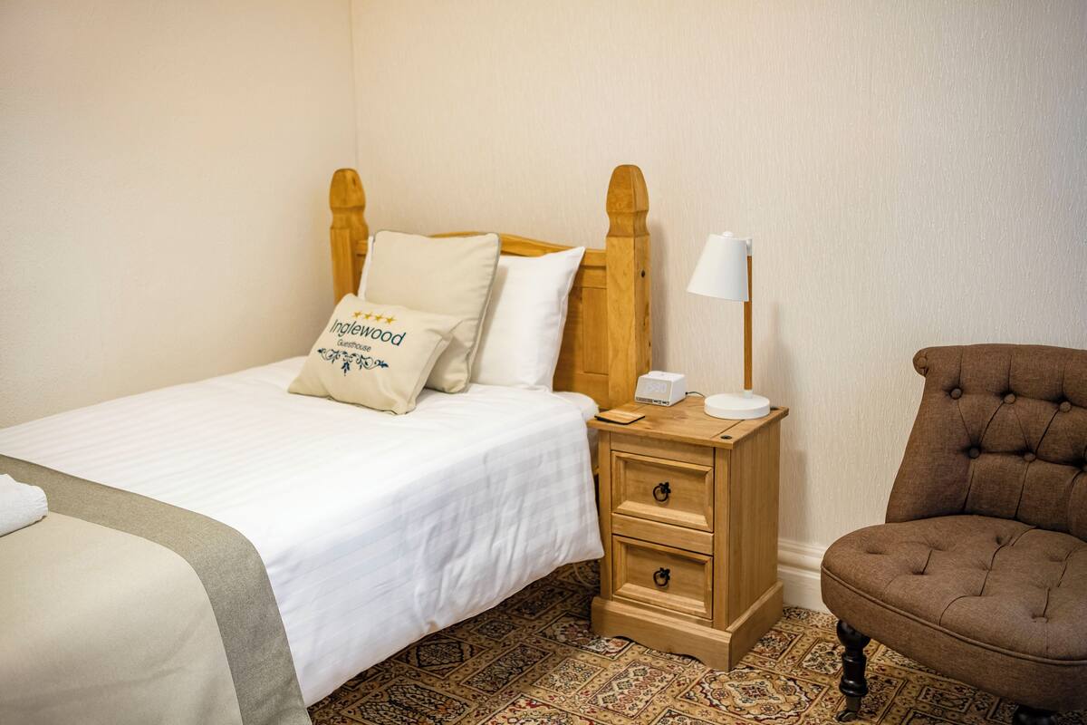A neatly made bed with a pillow, a bedside table with a lamp, a chair, and a patterned carpet.