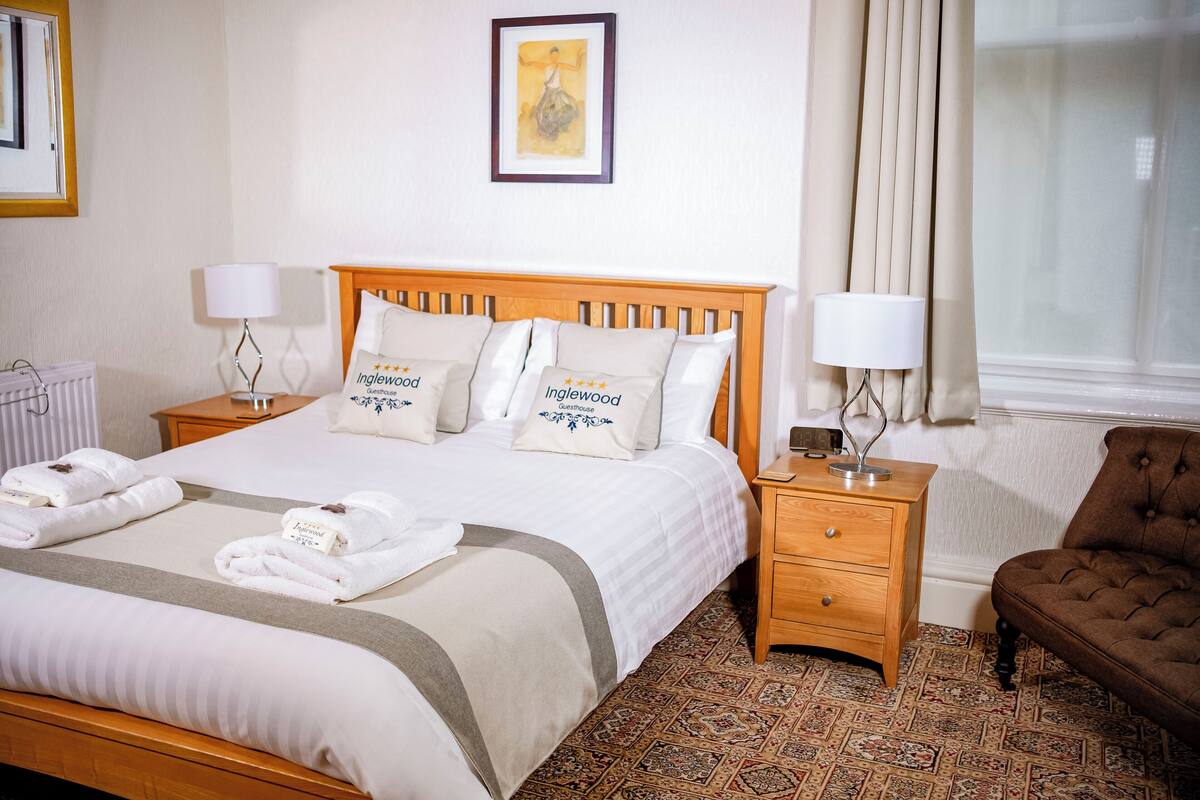 A neatly made bed with a wooden headboard, two bedside tables with lamps, a brown armchair, and a patterned carpet.