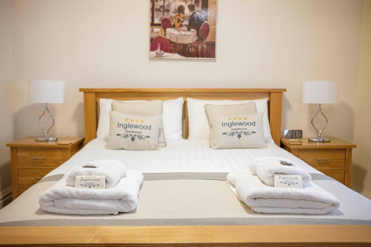 A neatly made double bed with two pillows labeled "Inglewood Guesthouse," two bedside lamps, and a folded towel on each side.