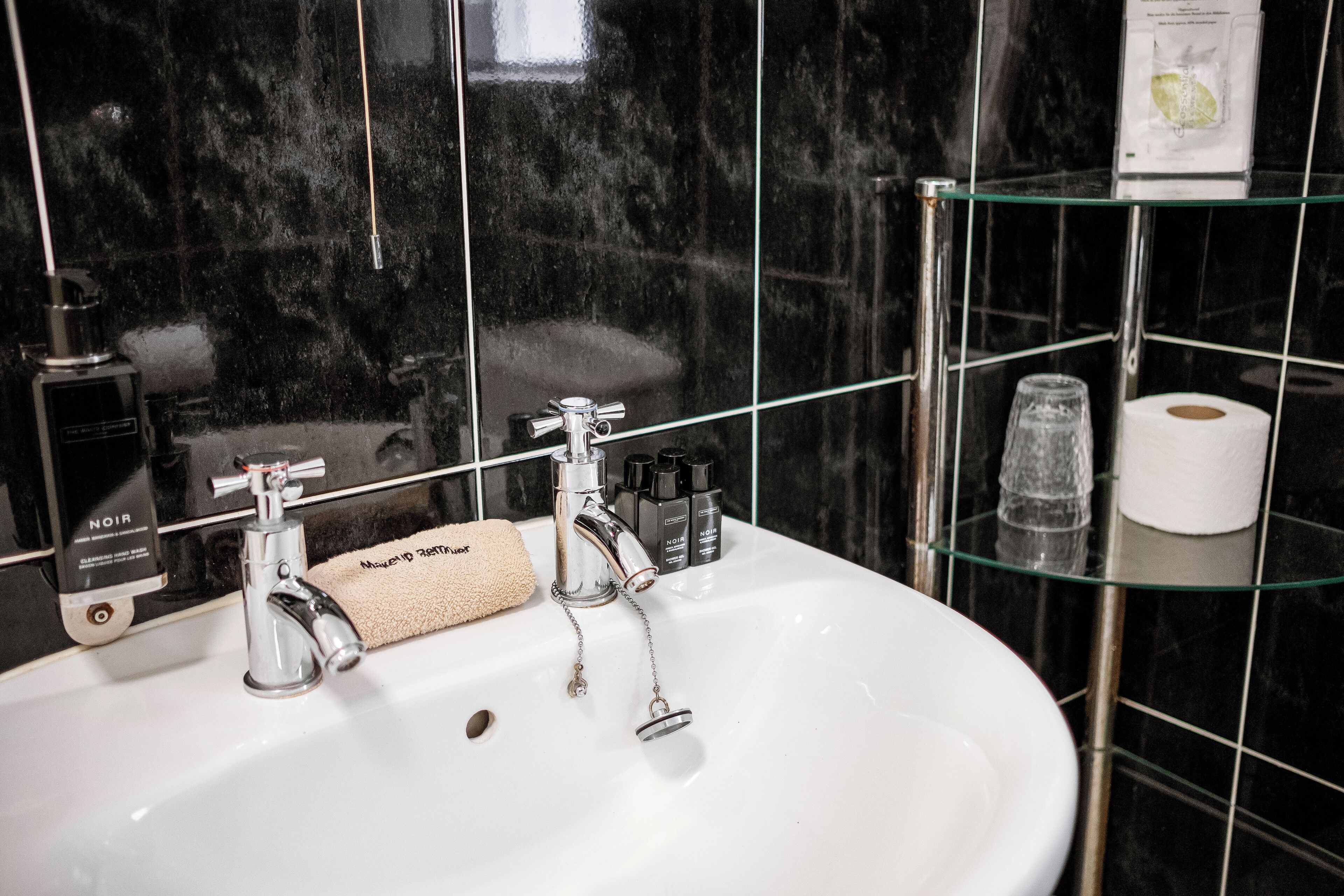 Ein Badezimmer mit einem weißen Waschbecken, zwei Wasserhähnen, einer Glasablage mit Toilettenartikeln und einer schwarzen Wand aus Fliesen.