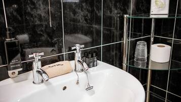 Ein Badezimmer mit einem weißen Waschbecken, zwei Wasserhähnen, einer Glasablage mit Toilettenartikeln und einer schwarzen Wand aus Fliesen.