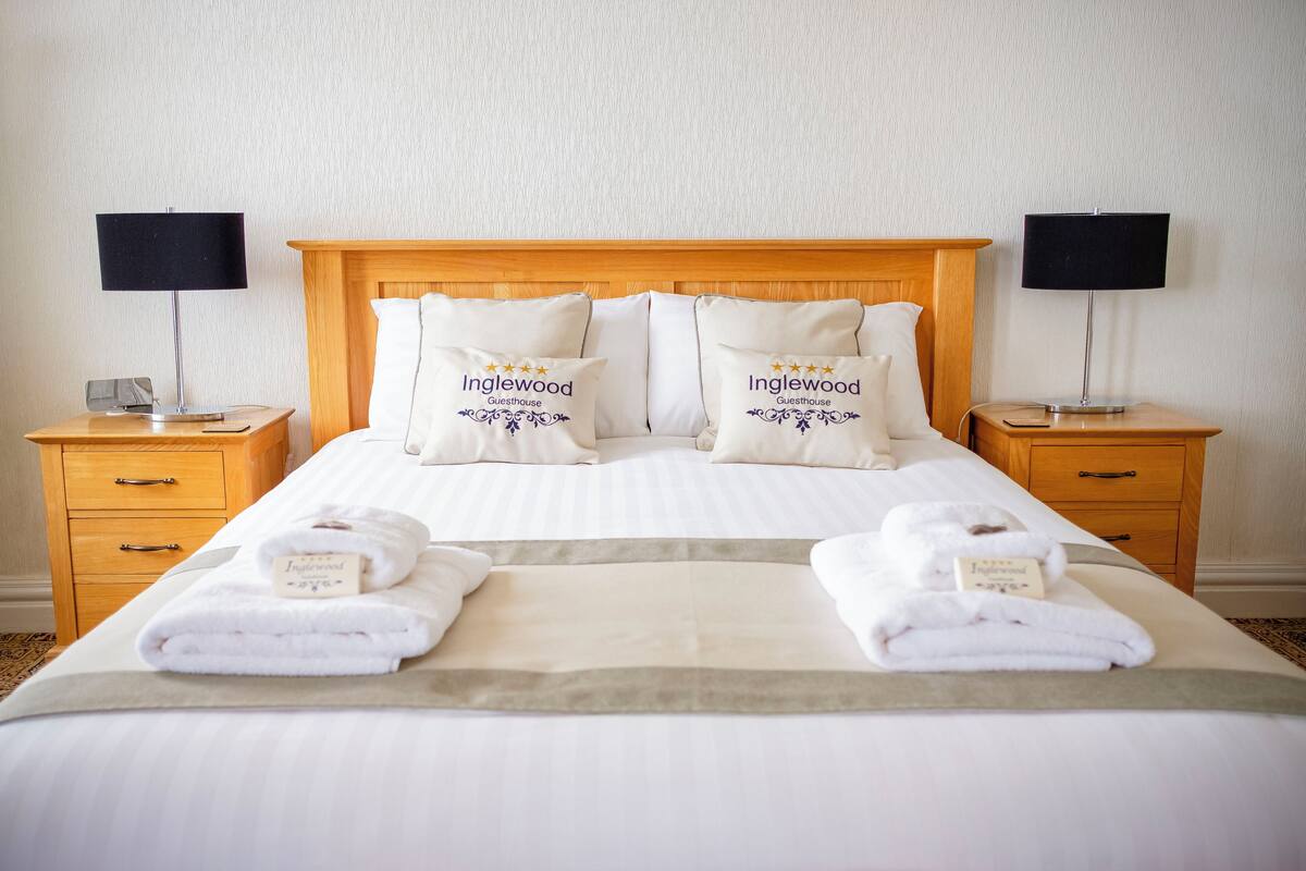 A neatly made bed with white linens, two bedside tables with lamps, and pillows with the logo "Inglewood Guesthouse".