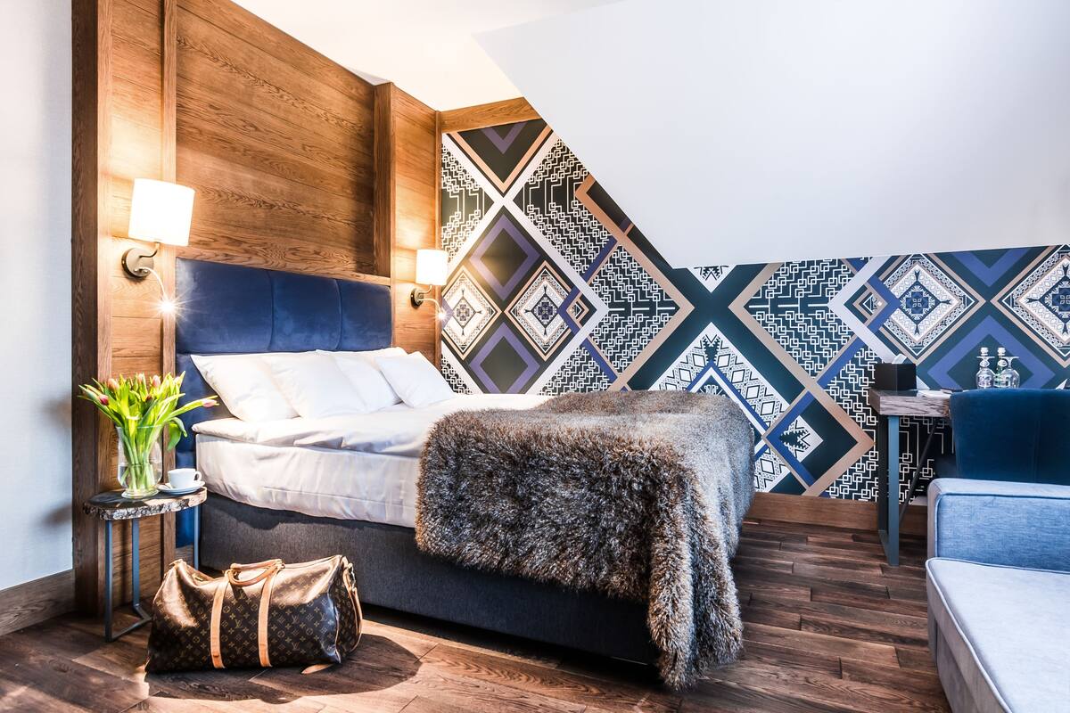 Een moderne slaapkamer met een houten hoofdbord, een bed met witte lakens, een zachte plaid, een Louis Vuitton tas en een behang met patroon.