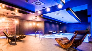 A modern indoor pool area with a wooden deck, lounge chairs, and a hot tub.