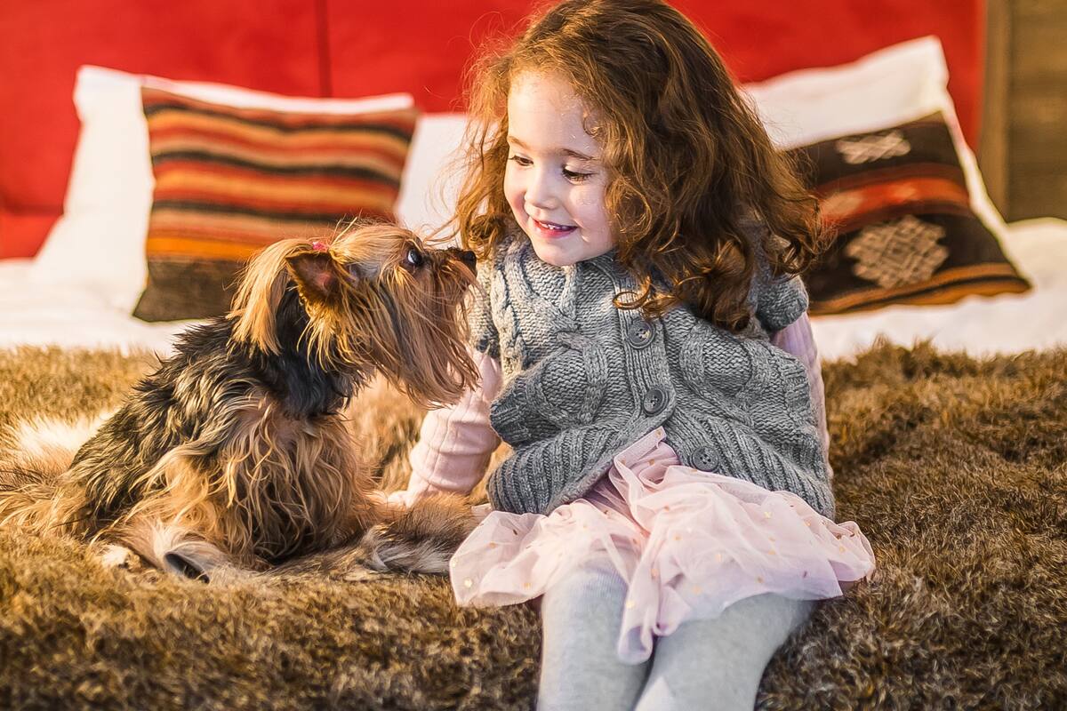 Een kind zit op een zachte vloerkleed met een hond, in een kamer met een rode bekleden hoofdeinde en gestreepte kussens.
