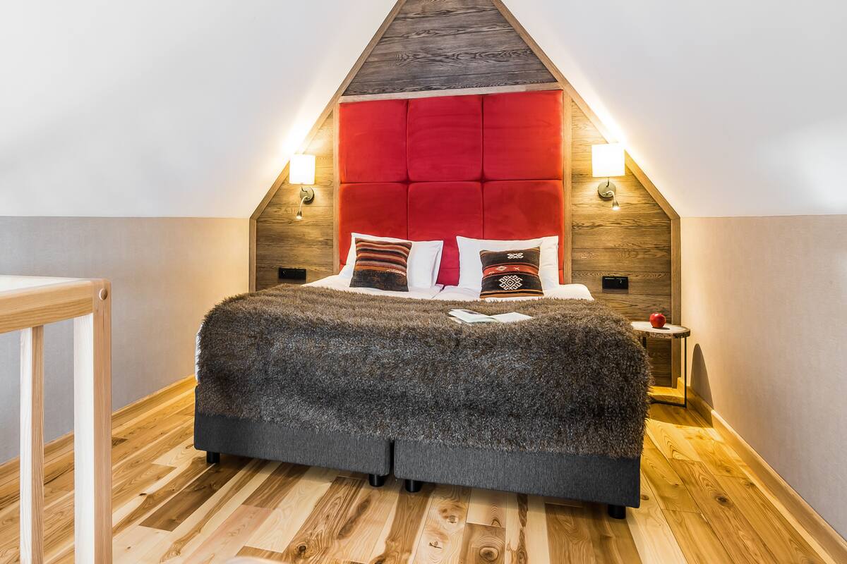 Een slaapkamer met een schuine balkenkap, een rode bekleden hoofdbord, een pluizige grijze sprei en twee bedlampjes.