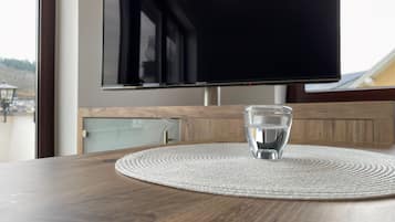 Een houten tafel met een glas water en een geweven placemat. Een flatscreen televisie is aan de muur boven de tafel bevestigd.