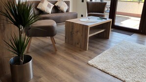 En moderne stue med et træsofabord, en sofa med puder, en stueplante og udsigt til en balkon med gelænder.