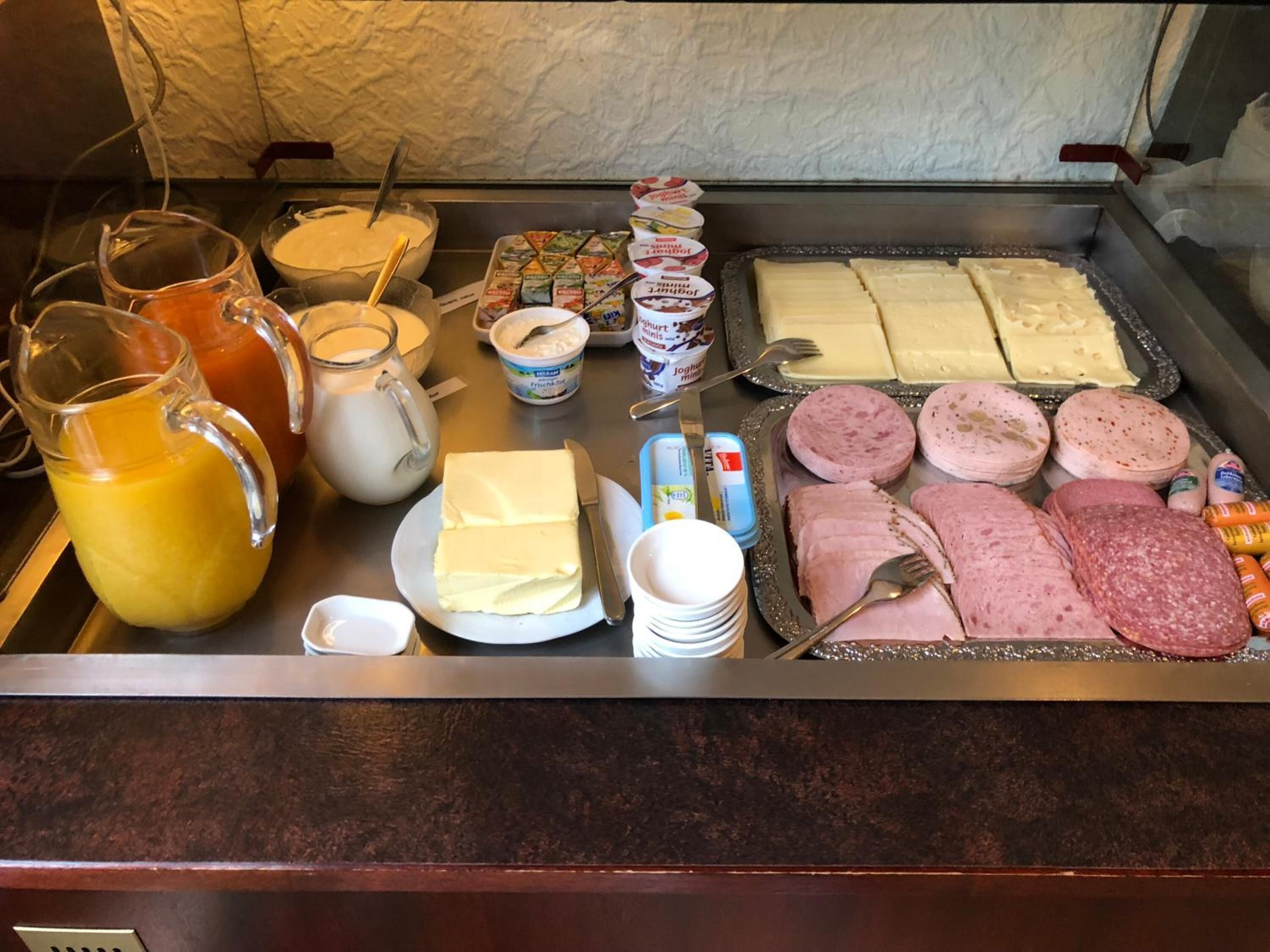 A breakfast buffet with various food items including butter, cheese, ham, sausages, and juices.