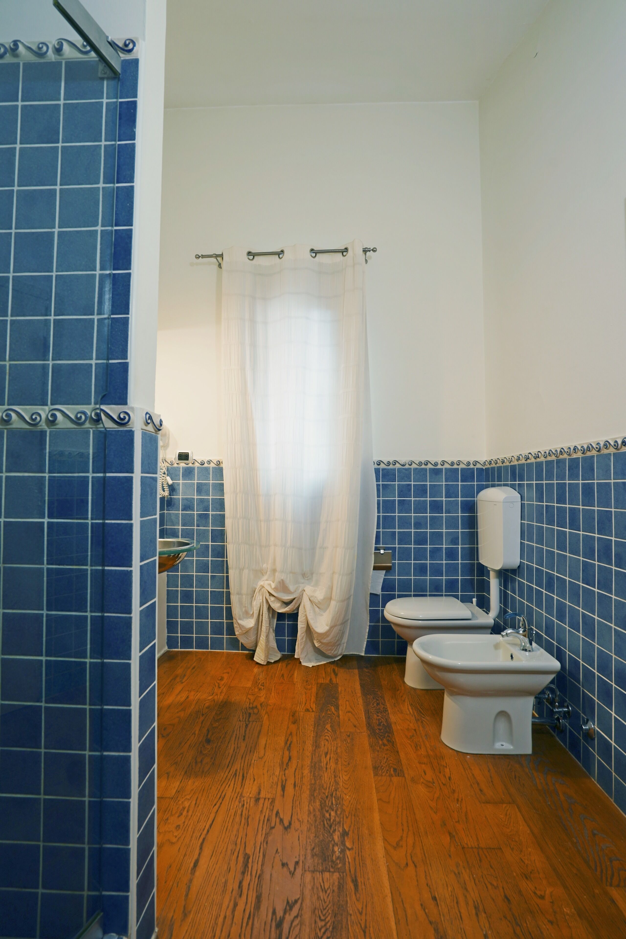 A bathroom with blue tiles, a wooden floor, a toilet, a bidet, and a shower curtain.