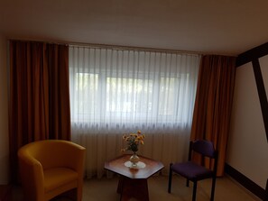 A room with a large window covered by sheer white curtains, a wooden table with a vase of yellow flowers, and two chairs, one yellow and one purple. - Landhotel Trampe (Breydin)