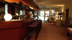A bar area with a long counter and multiple stools, a large round light fixture, and a view into a dining area with a table and chairs. - Landhotel Trampe (Breydin)