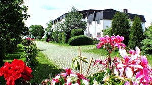 A large, white building with a black roof, surrounded by lush greenery and flowering plants. - Landhotel Trampe (Breydin)