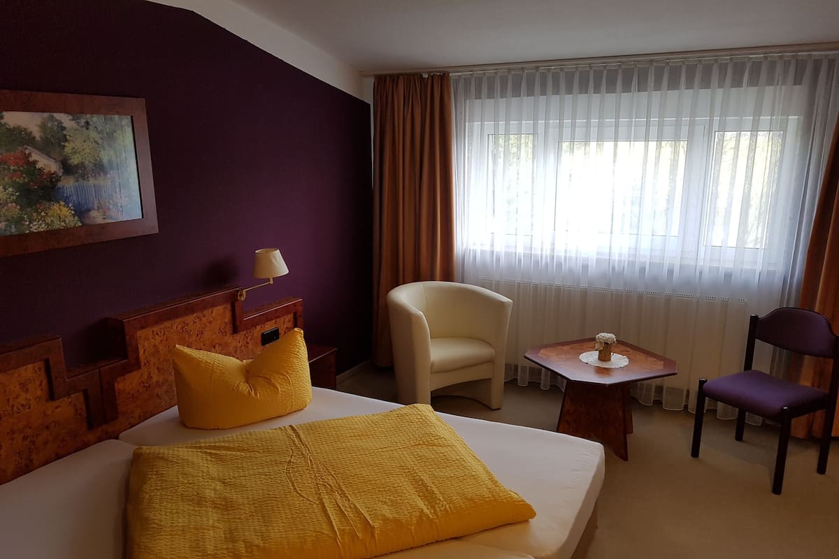 A hotel room with a bed, a chair, a small table, a lamp, and a painting on the wall.