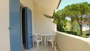A balcony with a table and two chairs, a blue door, and a view of trees and a house. - Flat in Lignano Riviera (Lignano Sabbiadoro)