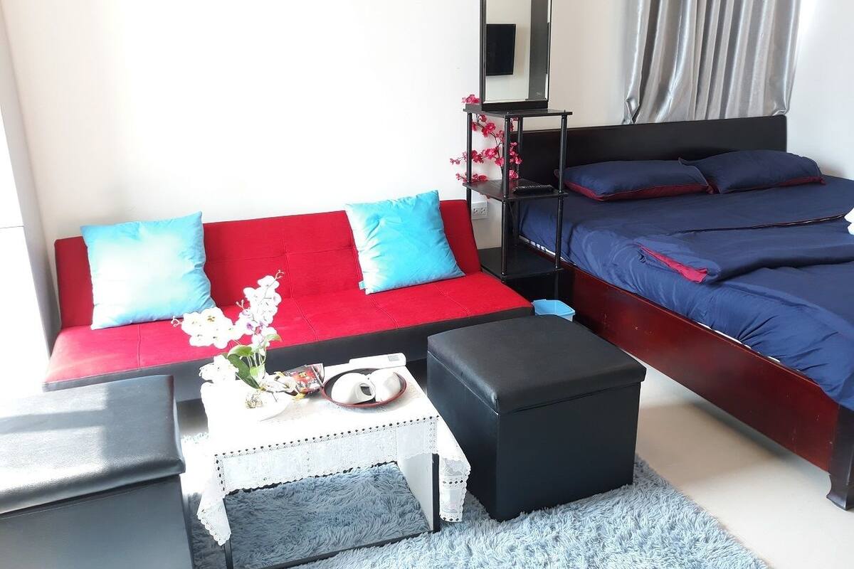 Une chambre d’hôtel avec un canapé rouge, un lit avec une literie bleue, une petite table blanche sur laquelle est posé un vase de fleurs, et un pouf noir.