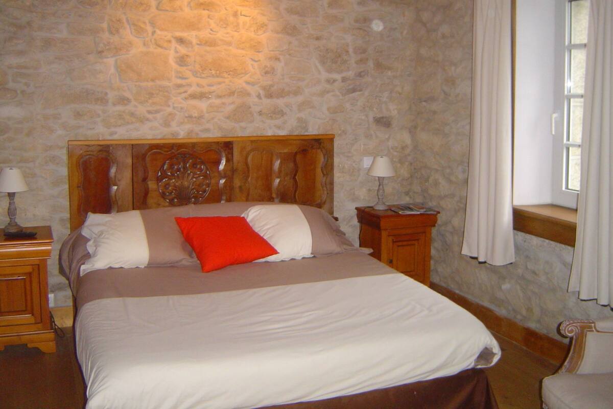 Une chambre avec un mur en pierre, une tête de lit en bois, un lit avec une literie blanche et marron, deux tables de chevet avec des lampes et une fenêtre avec des rideaux.