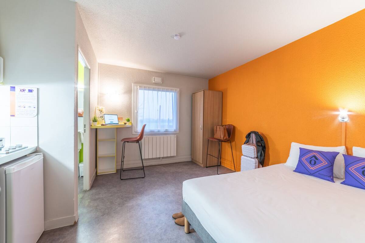 Quarto de hotel com cama, escrivaninha com computador, cadeira, guarda-roupa e radiador.
