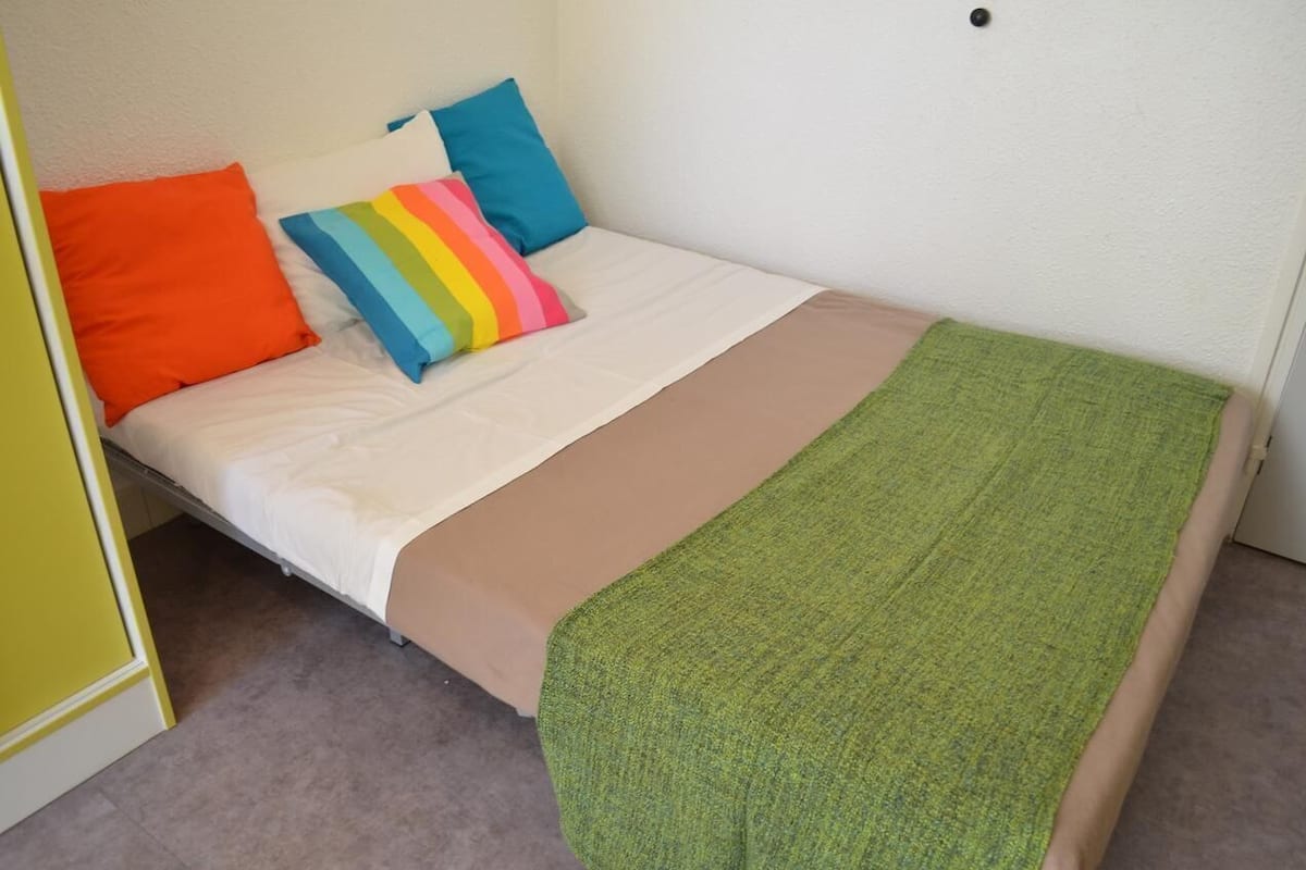 Uma cama de solteiro com colchão branco, manta verde e almofadas coloridas.