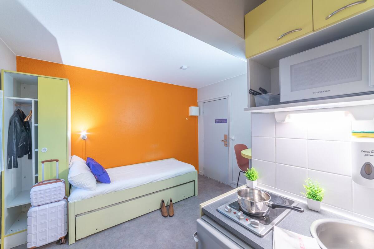 Quarto de hotel pequeno, com cama, guarda-roupa, cozinha compacta e banheiro.