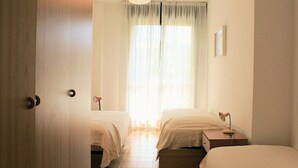 A hotel room with two beds, a wooden door, a bedside table with a lamp, and a window with sheer curtains.