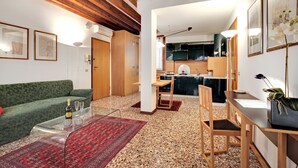 Ein Wohnzimmer mit einem grünen Sofa, einem Glastisch, einem roten Teppich, einem Holzesstisch mit Stühlen und einer Küche im Hintergrund.