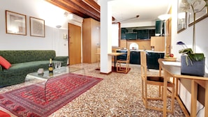 Ein modernes Wohnzimmer mit einem grünen Sofa, einem Glastisch und einem roten Teppich. Es gibt eine Küchenzeile mit Holzschränken und einem Esstisch mit Stühlen.