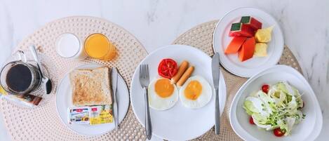Un desayuno con tostadas, huevos, salchichas, una ensalada de frutas y una bebida de café sobre una mesa de mármol.