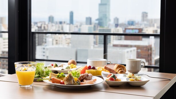 A breakfast table with various dishes and a glass of orange juice, set against a cityscape view through a window. - The OneFive Osaka Namba Kuromon (Osaka)