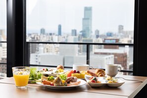 Una mesa de desayuno con diversos platos y un vaso de zumo de naranja, con vistas a un paisaje urbano a través de una ventana.