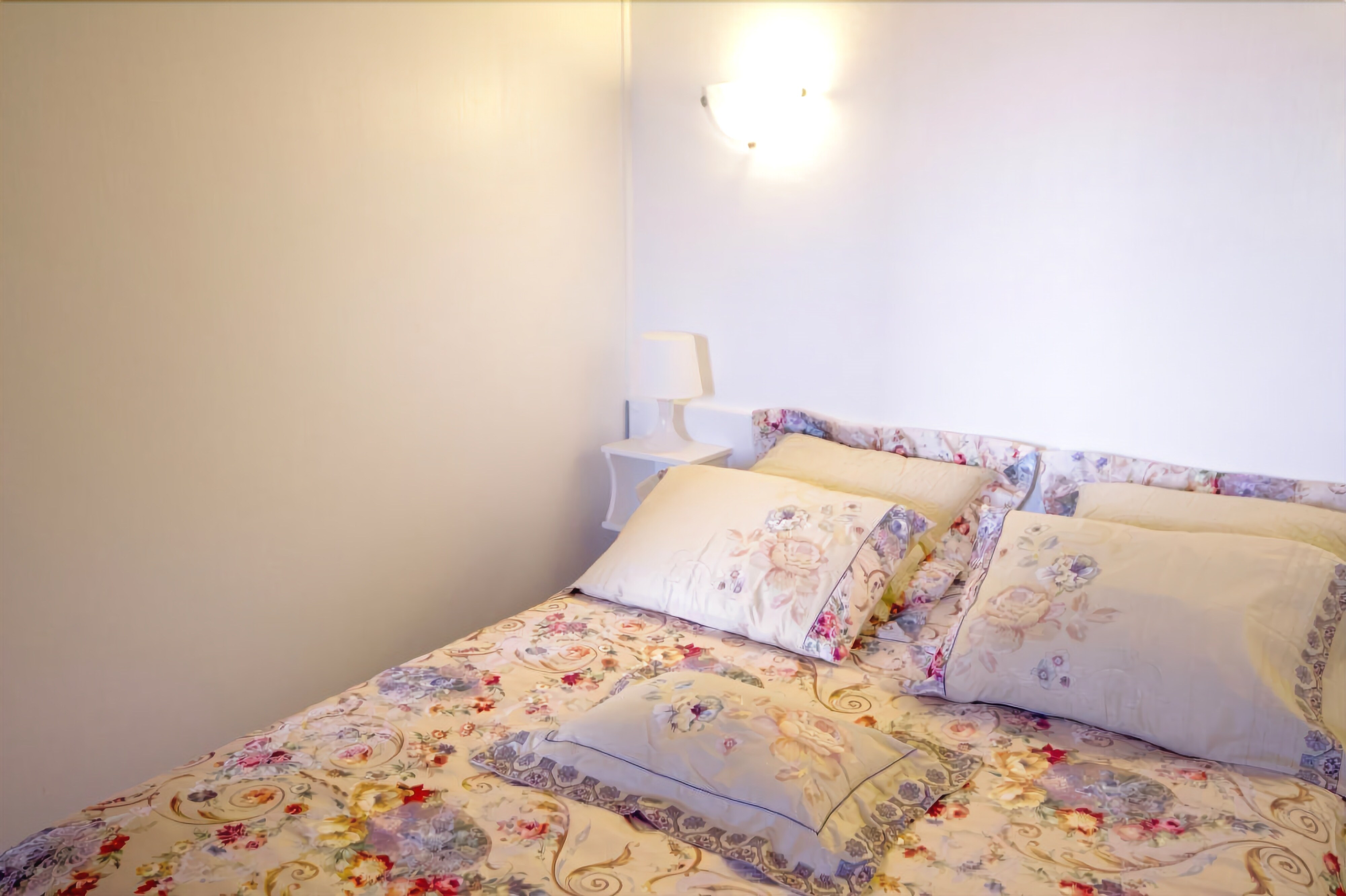 A neatly made bed with floral bedding and multiple pillows, a bedside table with a lamp, and a wall-mounted light fixture.