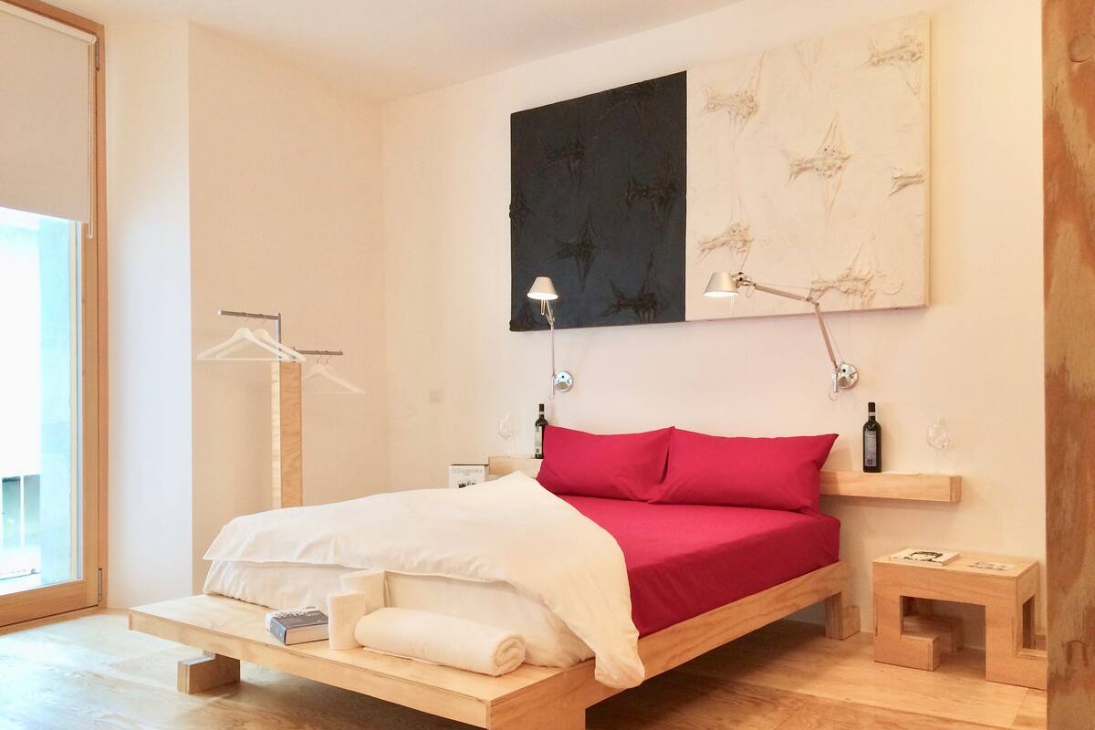 Un dormitorio moderno con una cama de madera, una almohada decorativa roja, una mesita de noche con lámpara y una pequeña mesita auxiliar de madera.
