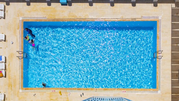 Et rektangulært swimmingpool med klart, blåt vand, omgivet af hvide liggestole og en fliseterrasse.