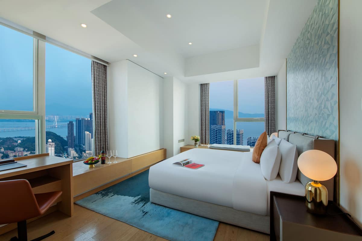 A modern hotel room with a large bed, a desk, and a view of the cityscape.