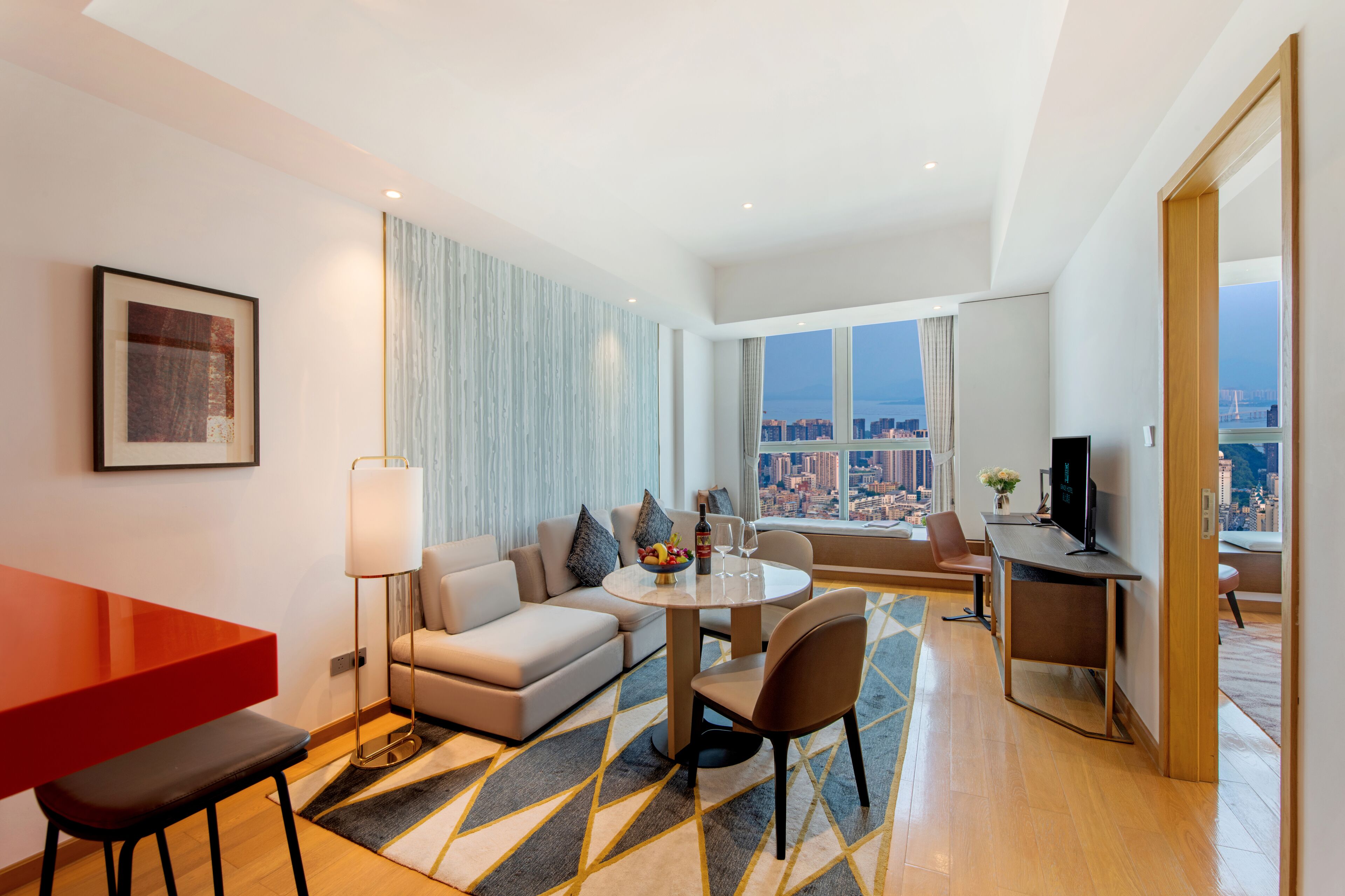 Habitación de hotel moderna con vista a la ciudad, zona de comedor, sofá, escritorio y televisión.
