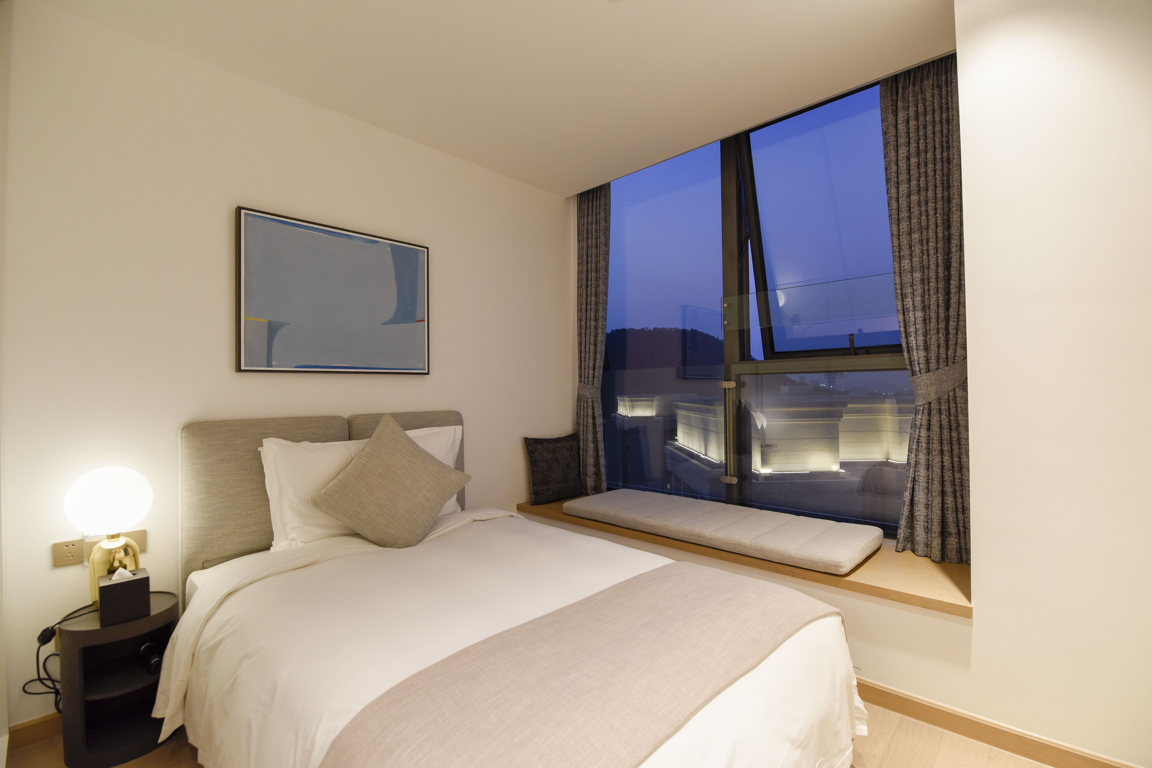 A hotel room with a large bed, bedside table, and a window with a view of the cityscape.
