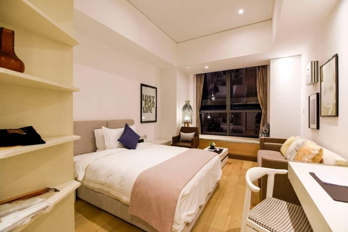 A hotel room with a bed, a desk, a chair, a window with a view, and a shelf with a vase.