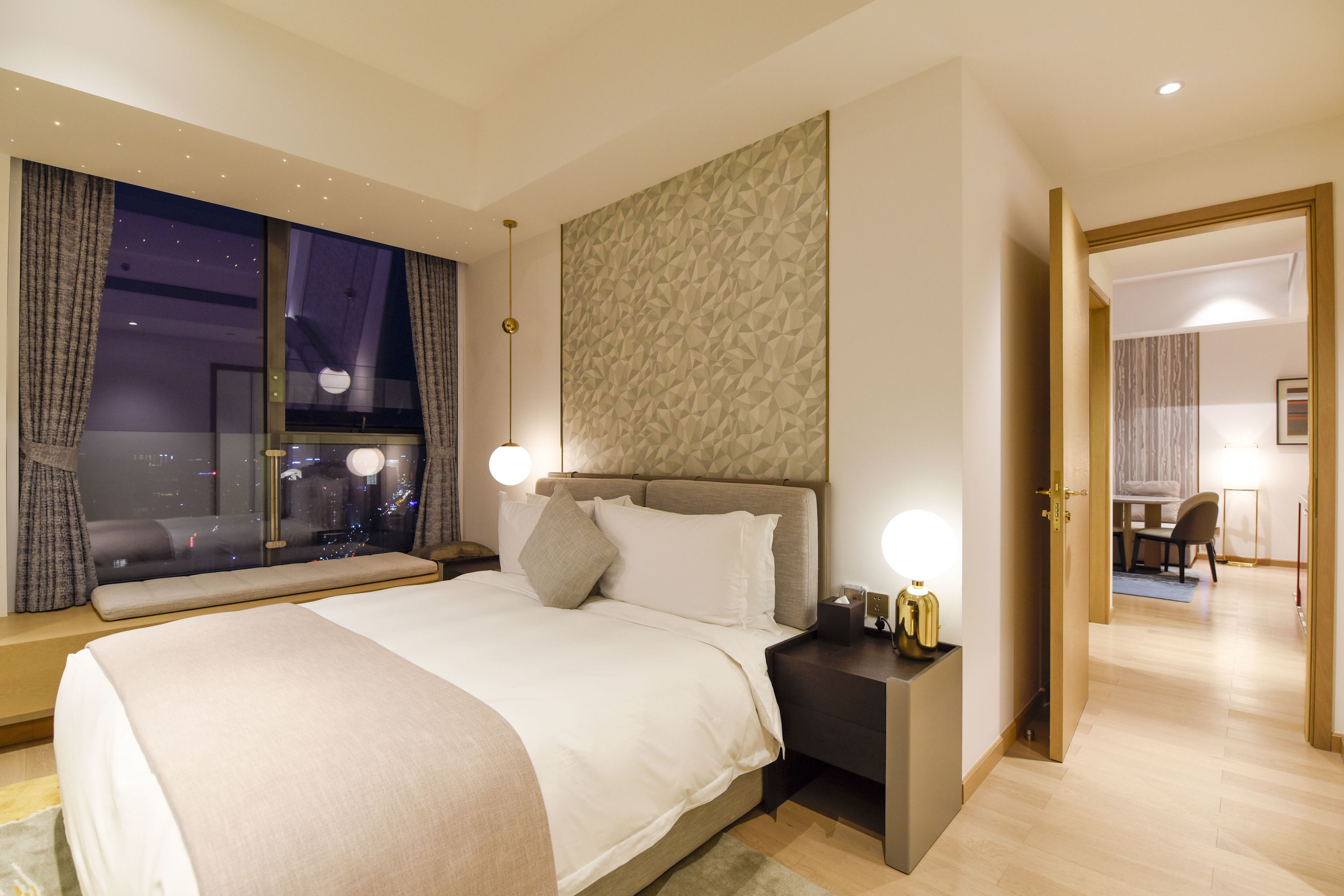 A modern hotel room with a large bed, a nightstand with a lamp, a view of the city through large windows, and a geometric wall design.