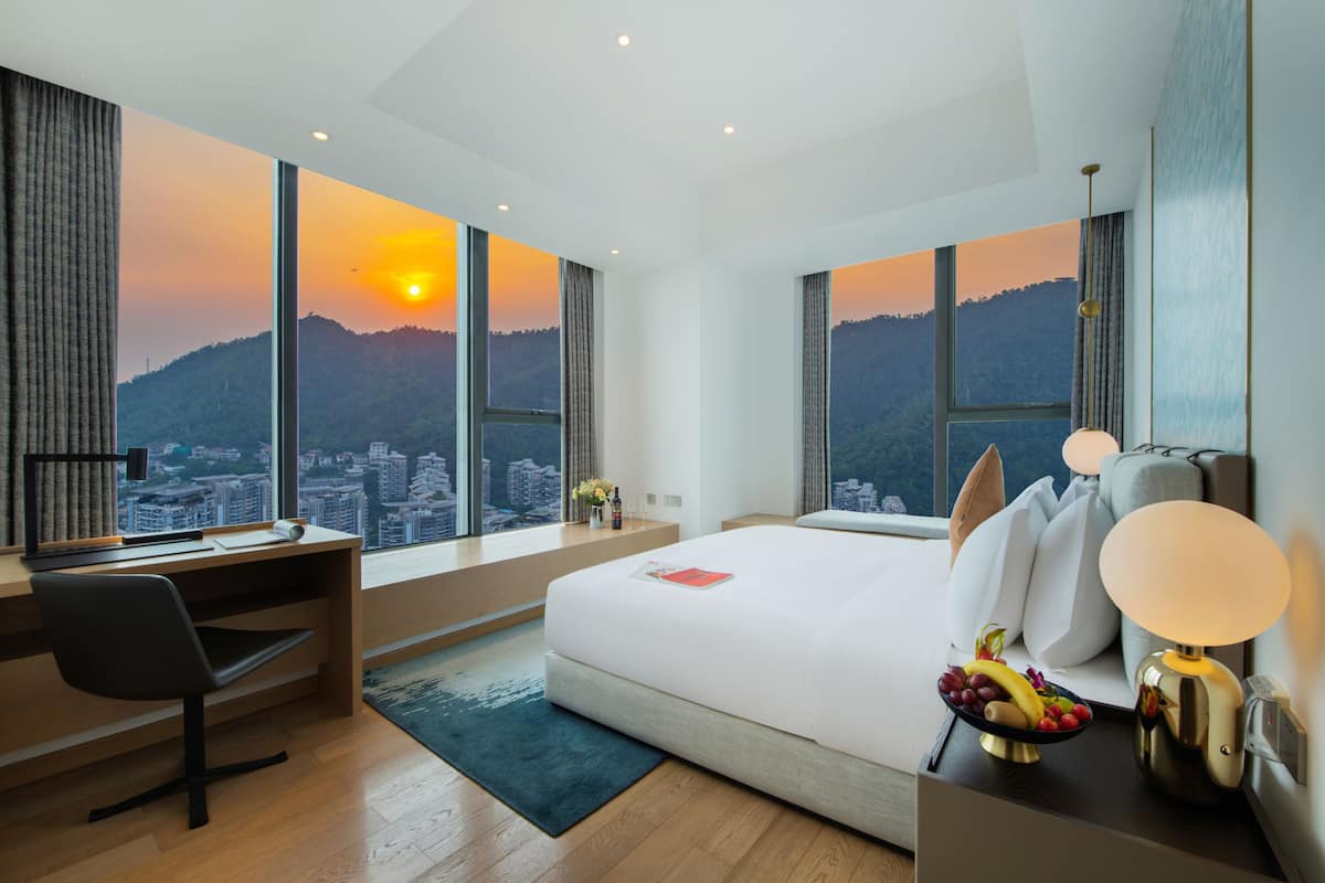 A modern hotel room with a large bed, a desk, and a view of a cityscape and mountains.