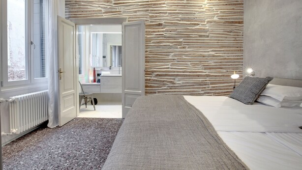 A modern bedroom with a stone wall, a bed with white linens, a bedside table, and a window with curtains.