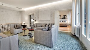 A modern hotel room with a living area featuring a sofa, armchairs, and a coffee table. There is a large window with a view of the city.