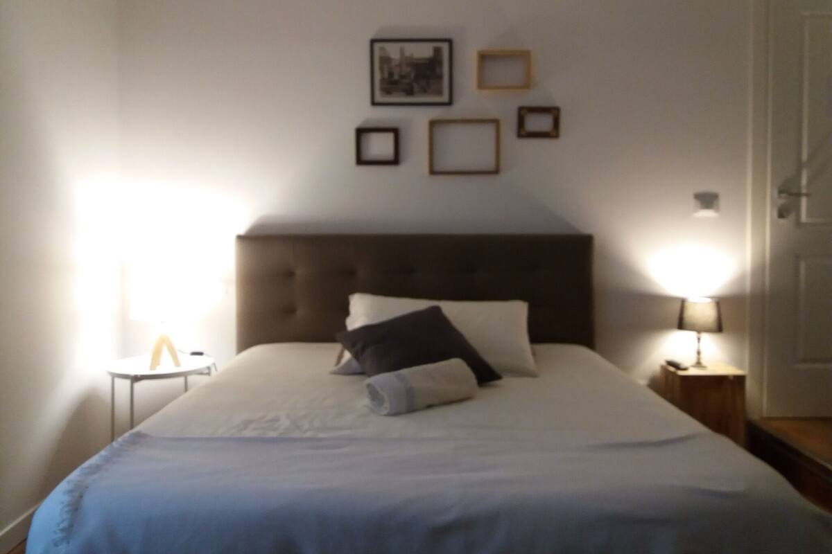 A neatly made bed with a headboard, a bedside table with a lamp, and framed pictures on the wall.