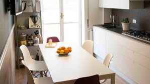 A modern kitchen with white cabinets, a dining table with chairs, a hanging light fixture, and a view of the outside through a door. - St. Peter's Luxury Rooms (Rome)