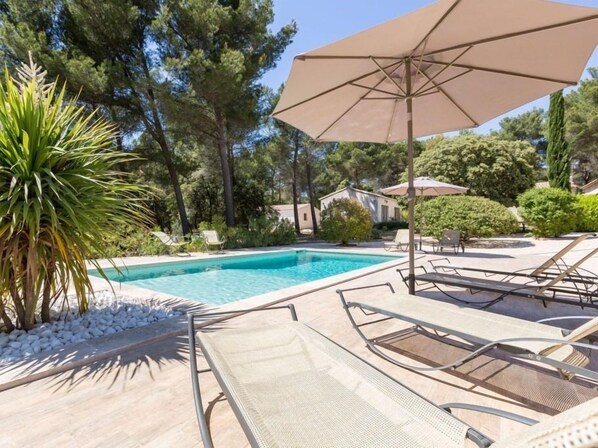 Una piscina con sillones de descanso y sombrillas, rodeada de árboles y un edificio pequeño.