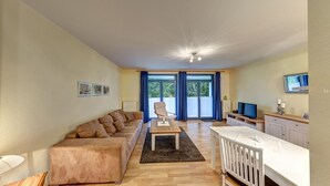 A modern living room with a sofa, a coffee table, and a TV. There is a large window with curtains, a door leading outside, and a small kitchen area with a TV and a cabinet.