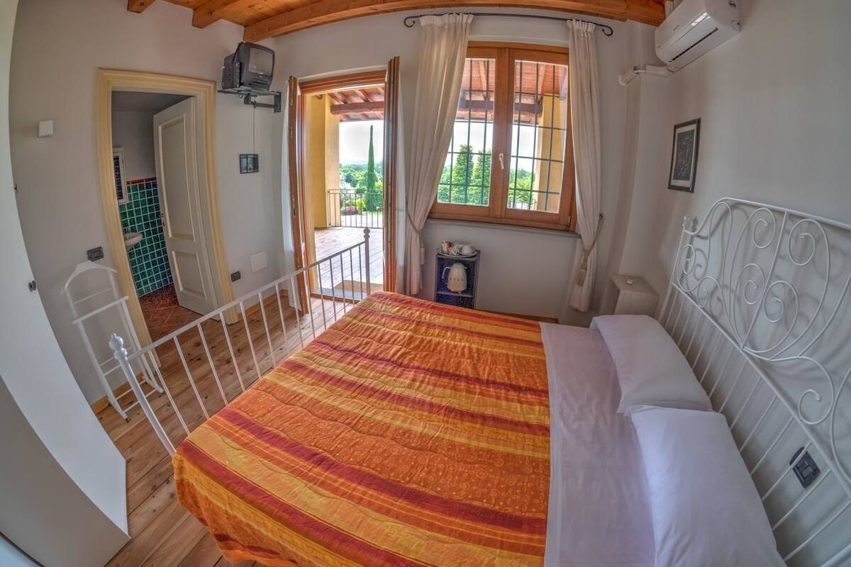 Ein Schlafzimmer mit Bett, einem Fenster mit Vorhängen, einem Balkon und Blick ins Grüne.