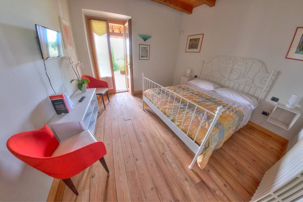 Ein Schlafzimmer mit Holzboden, einem Bett mit gemustertem Überwurf, einem roten Sessel, einem Schreibtisch mit Computer und einem an der Wand befestigten Fernseher.