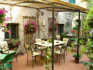 Une terrasse avec une table et des chaises, entourée de plantes en pot et de fleurs suspendues.