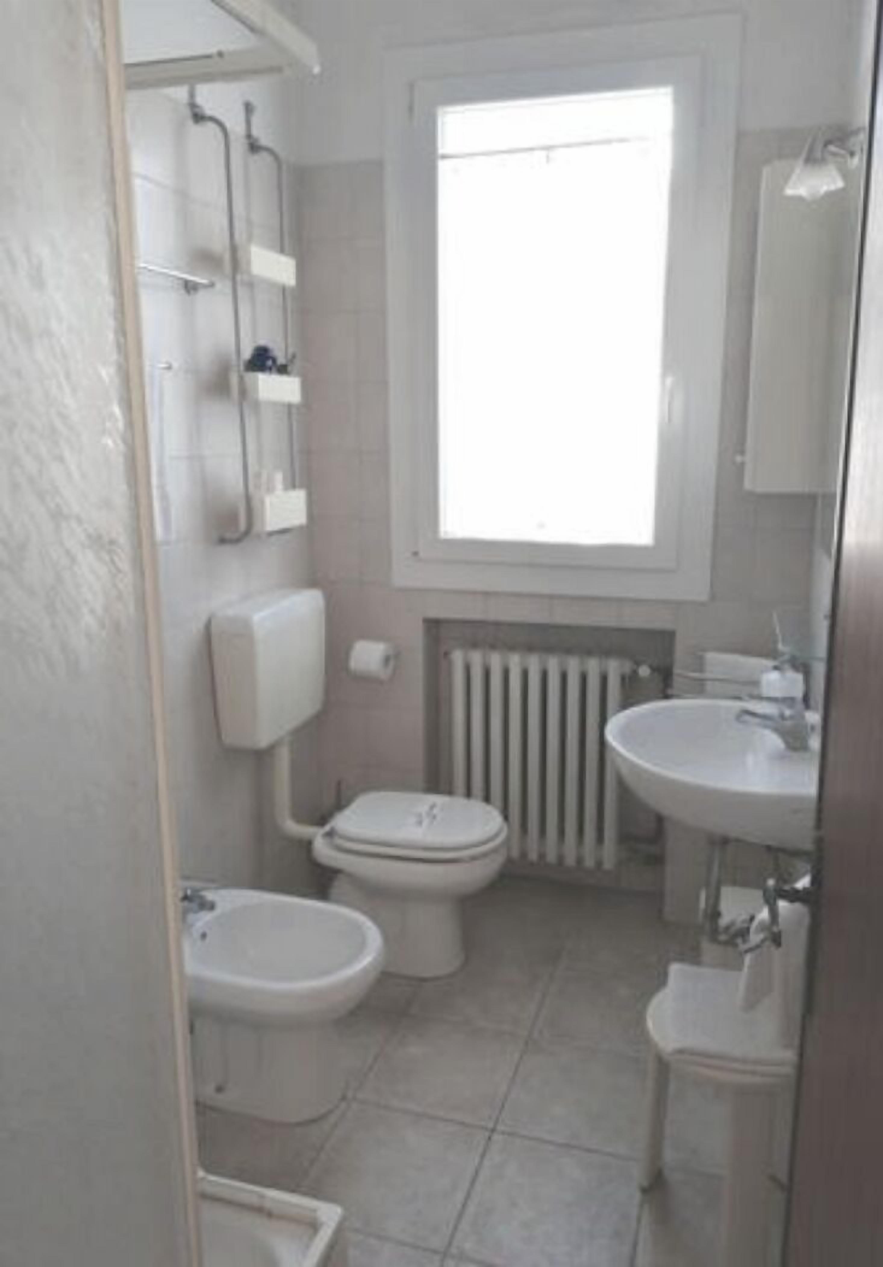 Baño pequeño y funcional con inodoro, bidé y lavamanos. Una ventana aporta luz natural y hay un radiador debajo.
