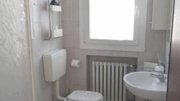 Baño pequeño y funcional con inodoro, bidé y lavamanos. Una ventana aporta luz natural y hay un radiador debajo.